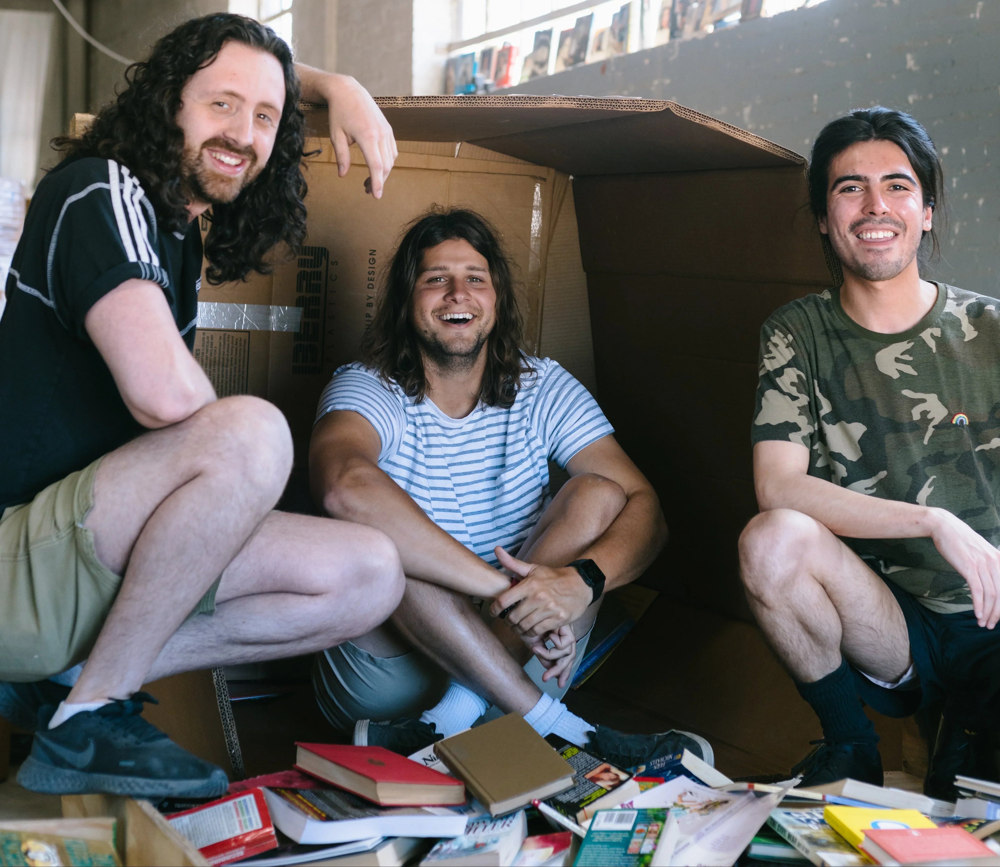 Three people sitting inside a cardboard box in a room with large windows.